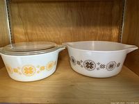 Photo showing both casserole dishes side by side on a wooden shelf. The yellow patterned smaller dish includes a lid.