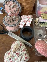 Photo of pincushions with pearl-headed pins, scissors, measuring tape, and small black ring-shaped item on wooden surface.