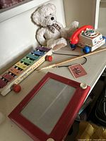 Vintage toys displayed on shelf including Fisher Price xylophone, rotary phone, Etch-A-Sketch, plush bear, and mallet