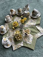 Top-down view of tea cups, saucers, cream pitcher, and porcelain yellow rose floral figurines arranged on fabric mat