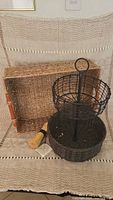 Rectangular woven tray, two-level black metal fruit basket, and straw potato scrubber displayed together on cloth.