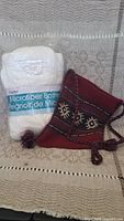 White microfiber bathrobe brand new in packaging next to a maroon handmade decorative bag with traditional Greek embroidery and braided tassel strap.