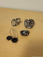 Four pieces of costume jewellery: three decorative silver-tone rings and one pair of black stone drop earrings arranged on a wooden surface.