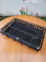 Photo of a folded black metal dog crate with metal bars and plastic tray, placed on a wooden surface outdoors with flowers in the background.
