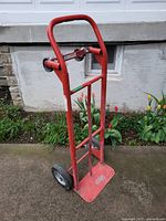 Front and side view of a red dolly cart with two black solid wheels and a curved handlebar, positioned outdoors on a concrete surface with flowers in the background.