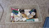 Plastic container with multiple tubes and bottles of various fabric paints and paint pens in colors including white, pink, blue, gold, teal, black, and yellow.