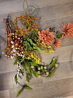 Assorted artificial flowers laid out on floor including orange daisies, yellow sprays, berry stems, and green foliage.