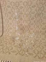 Full view of the clear glass decanter with stopper and six matching spirits glasses on a patterned fabric background.