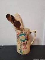Porcelain pitcher with floral designs holding several wooden kitchen utensils against a white wall.
