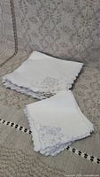 Stack of folded white linen napkins with purple embroidery on corner, showing floral and butterfly motifs, scalloped edges