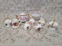 Full view of the miniature porcelain tea set pieces arranged on a patterned fabric background to show details of floral patterns and gold trim.