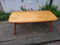 Rectangular wooden coffee table shown on a concrete surface outdoors with flowers in the background. The table is photographed from an angle showing the top surface and two turned legs on each side.