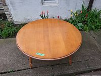 Round wooden coffee table shown from an angled top view outdoors with garden flowers in the background.