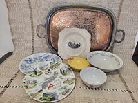 Photo of vintage servingware including a large rectangular metal embossed tray, five decorative plates with various designs, and two ceramic bowls.