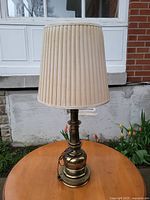 Front view of solid brass lamp on wooden table with pleated beige shade outdoors