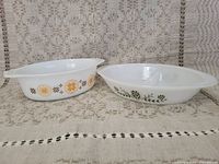 Two vintage Pyrex dishes shown from the side, one with a yellow floral design and one plain white with handles.