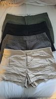 Four pairs of women's shorts in beige, gray, black, and olive green stacked flat showing front waistbands and button closures.