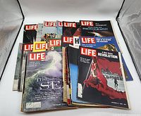 Collection of more than 25 vintage Life magazines arranged in a fan shape, showing colorful photographic covers including ocean wave and revolution scenes.