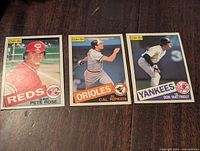 Three 1985 O-Pee-Chee baseball cards featuring Pete Rose, Cal Ripken, and Don Mattingly laid out on a wood surface.
