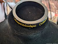 Black bangle bracelet showing Catherine Malandrino yellow logo text on black background, displayed on black stand.