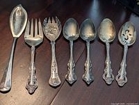 Top view of seven silver plated serving utensils showing varied spoon and fork shapes with detailed decorative handles.