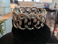 Silver tone cuff bracelet displayed on black stand, showing interlocking oval links with textured surfaces and overall structure.