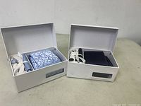 Both men's tie sets with cuffs displayed in their white boxes, showing one light blue swirl patterned tie and one solid dark tie with cufflinks.