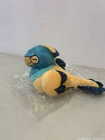 Side view of the blue and yellow-orange Shortcake Archaeopteryx plush on clear plastic wrapping.