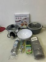 Photo showing electric pot with black base and glass lid, gray pot or saucepan with lid, white bowl, new acrylic cupcake stand box, steel wool scrubbers, Scotch-Brite scrubbing pad, and vegetable peeler arranged on table.