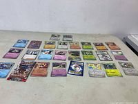 23 assorted Pokémon trading cards spread out on a flat surface, showing various characters and card types including holographic and regular cards.