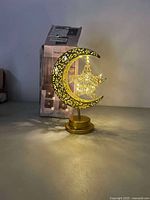 Moon and star neon light illuminated, with metal cut-out decorative pattern and circular base. Box and packaging visible behind the lamp.
