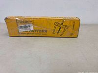 Yellow rectangular box with printed illustration of a two-arm puller tool and size information 127-315mm. Box is wrapped with tape and shows ware.