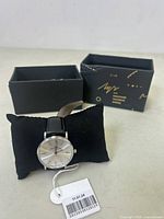 Photo showing the Minsk Sarga quartz watch on a black cushion with the original branded black and gold box in background. The watch has a silver dial and black leather strap with a white price tag.