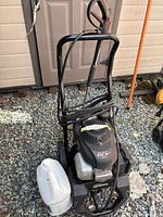 Overall front view of pressure washer on gravel showing metal frame, wheels, coiled hose, engine top, detergent jug beside unit