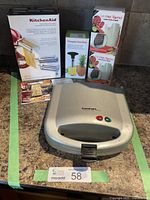 Overview of Cuisinart sandwich grill with accompanying boxed accessories: KitchenAid pasta attachment, pineapple corer, and vegetable slicers