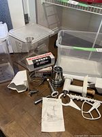 Photo of KitchenAid attachments including sausage stuffer parts, metal grinder pieces, a flat beater, instruction manual, and two large clear plastic mixing bins and storage bin.