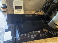 Top view of NutriChef dual induction cooktop on countertop with manual behind it, showing dual cooking zones and part of control panel.
