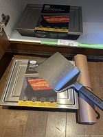 Photo showing two OONI Detroit-style pizza pans with box packaging, an OONI pizza paddle with black handle on top, and a rolled silicone baking mat.