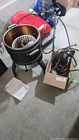 Overall view of large black metal outdoor gas deep fryer with perforated frying basket inside, gas accessory components, and instruction manual on floor beside the fryer.
