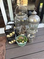 Photo showing two large glass jar lanterns with metal handles, a smaller glass jar, a metal lantern, three boxed solar glass decoration lights, and a small potted succulent plant on outdoor deck.