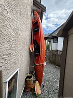 Front angled view of red-orange Wilderness Systems Pamlico 135T kayak leaning against exterior wall with two paddles strapped