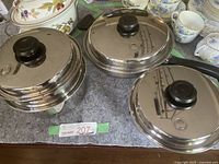 Three lidded stainless steel cookware pieces arranged on a protective pad with china dinnerware in background.