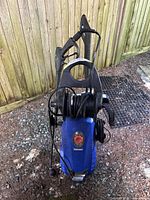 Front and angled views of blue Simoniz S1900 electric pressure washer with black hose reel, spray gun, and wand, outside on gravel