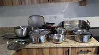 Cookware assortment on countertop showing frypans, griddle pan, large roaster, pots with lids, and stainless bowls.
