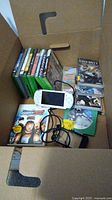 Open cardboard box showing multiple Xbox and PSP game cases and a white Sony PSP handheld console lying flat with charging cable inside the box