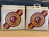Four vintage white vinyl stadium seat cushions with burgundy and gold Washington Redskins helmet logo and diagonal stripes, two with handles.