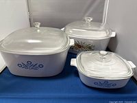 Photo showing all three CorningWare casserole dishes with glass lids on a blue surface, two with Blue Cornflower pattern and one with Spice of Life pattern