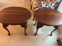 View of two oval brown wooden end tables side-by-side on carpet near patterned chair, highlighting shape and legs.