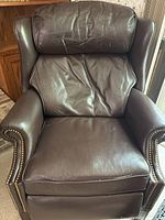 Front view of the brown leather recliner chair showing creased leather on the backrest and seat with brass nailhead trim along the arms.