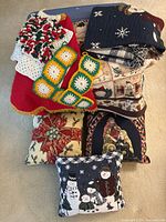 Grouped holiday-themed textiles shown from above: pillows with snowmen, nativity, bow, and houses; blankets including navy blue with snowflakes and red fleece with crocheted wreath and patches.